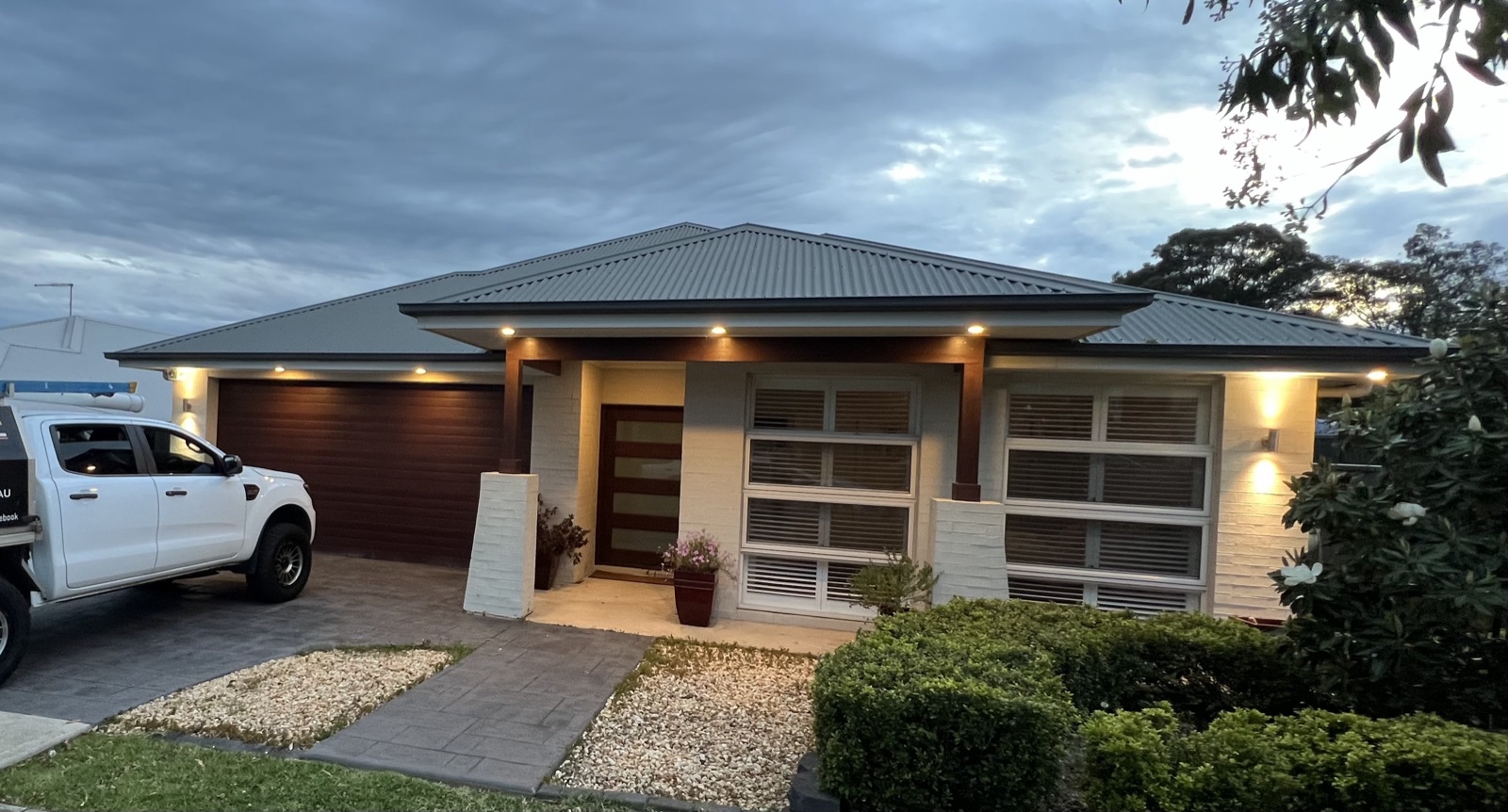 Single-storey Sydney home at dusk with warm soffit downlights under the eaves and uplit entry pillars — Power Play ute in the driveway